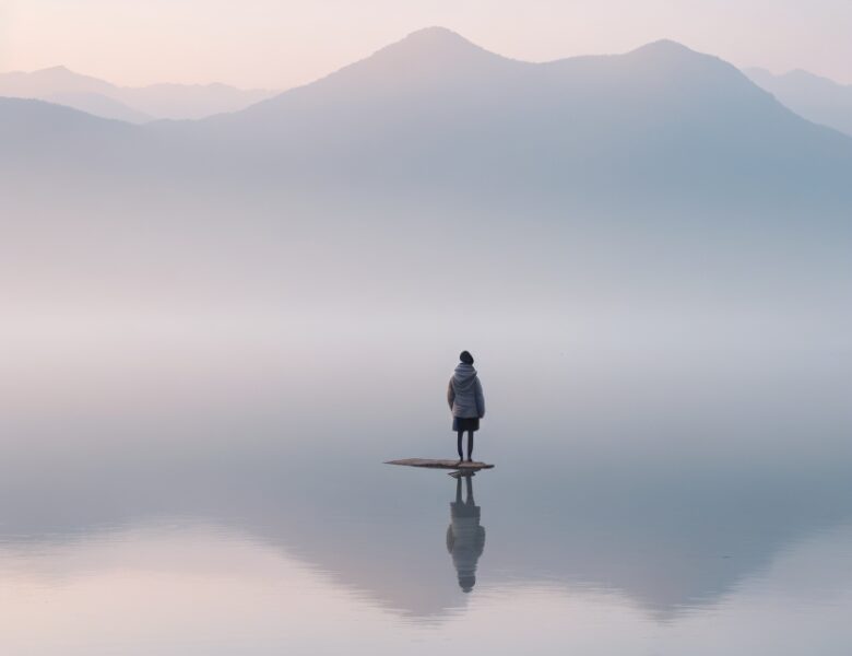 Einzelperson am Rande eines Sees blickt auf Berge, Beziehung zwischen Mensch und Natur.
